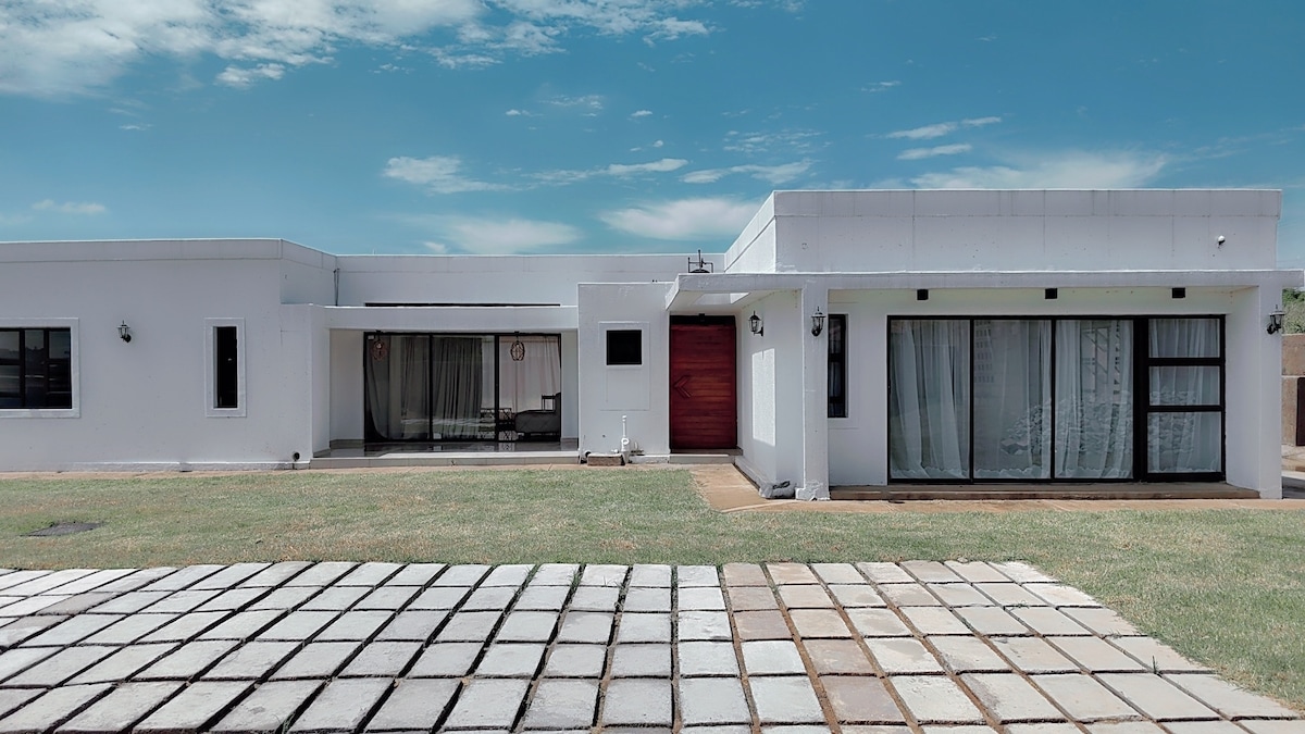 The exterior of a modern one-story home is presented with a large, grassy yard. Glass doors provide a view into the interior, while a wooden front door adds a contrasting accent. A spacious patio area features a stone path leading to the entrance.