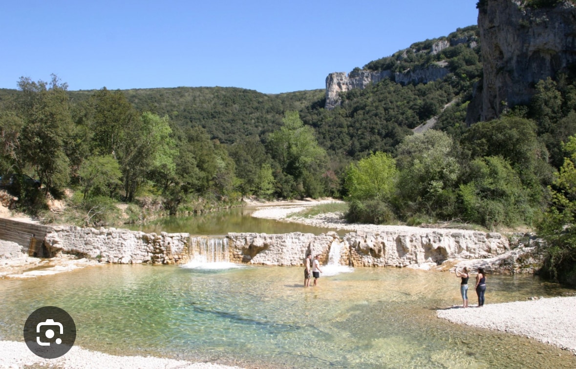 A serene river scene is depicted, with clear turquoise waters gently flowing over a rocky ledge. Lush greenery frames the landscape, and several people are seen enjoying the water on the riverbank. The sun is shining brightly in a clear blue sky.