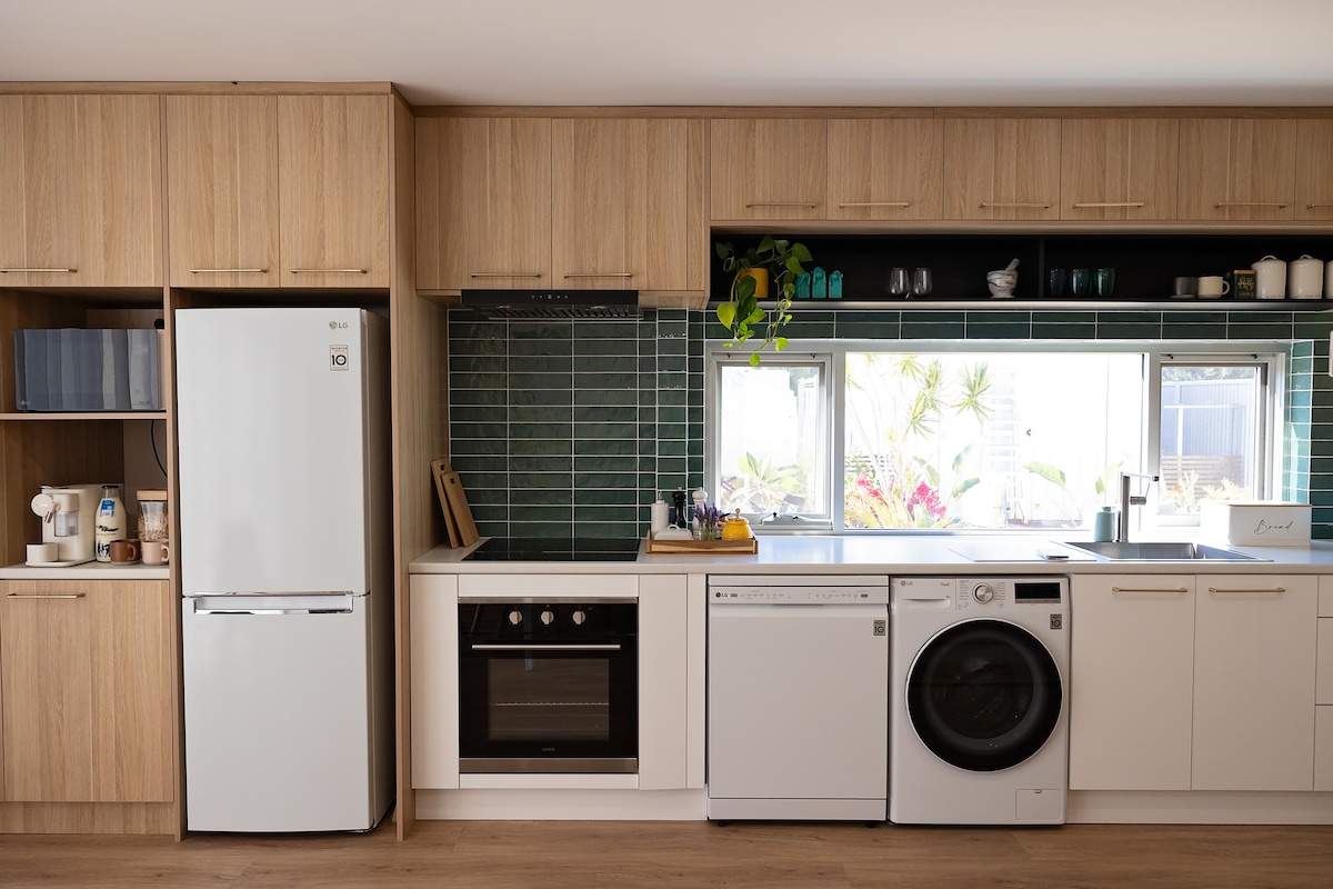 The kitchen features modern cabinetry and stainless steel appliances, including a refrigerator, oven, and dishwasher. A stylish sink is positioned beneath a window that provides natural light. A washing machine is also integrated into the cabinetry, creating an efficient and functional space.