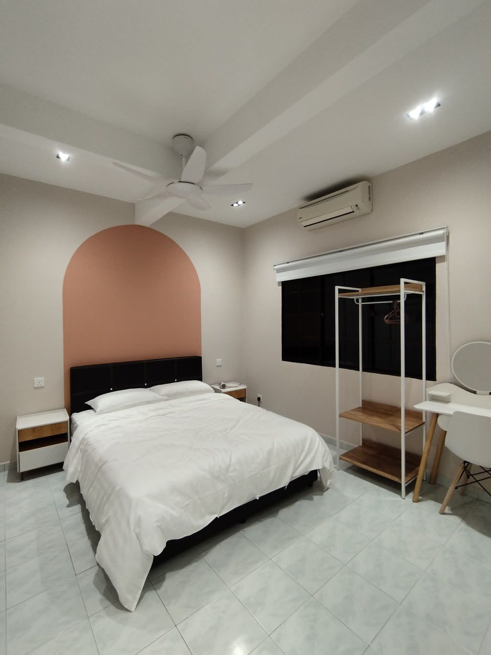 A well-lit bedroom is presented with neutral-colored walls, featuring a spacious bed with a white comforter. A decorative arch in a warm tone is positioned above the bed. The room includes a small wardrobe and a modern desk, with a ceiling fan providing airflow.