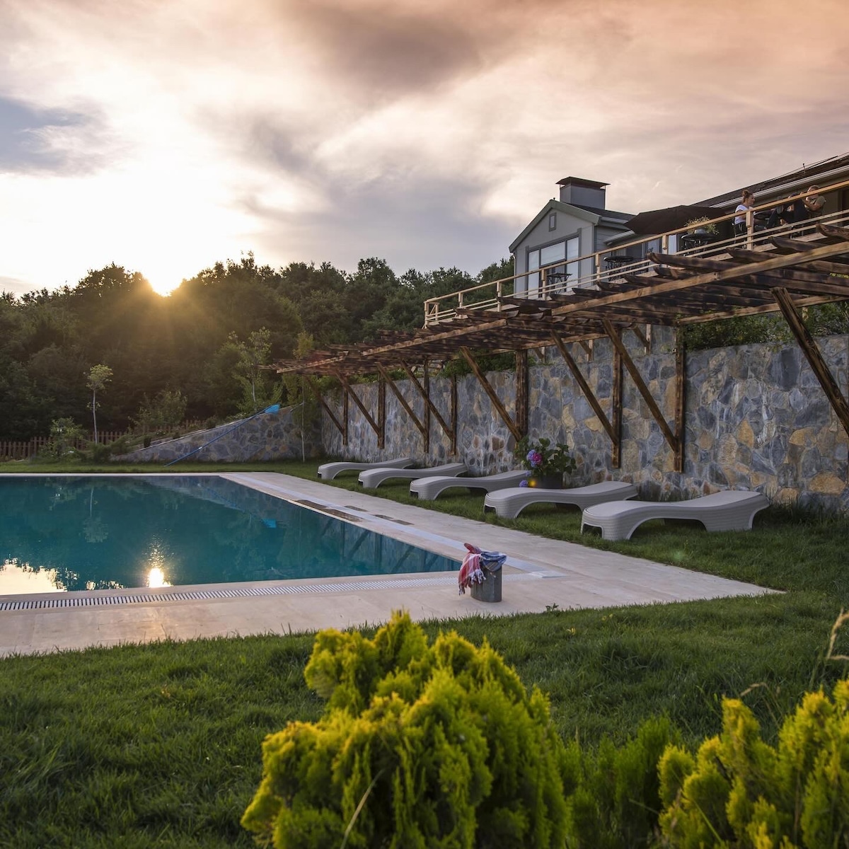 A tranquil pool area is framed by lush greenery and a wooden pergola. Sun loungers are positioned by the water's edge, with soft grass surrounding the space. The setting sun casts a warm glow over the picturesque landscape, enhancing the serene atmosphere.