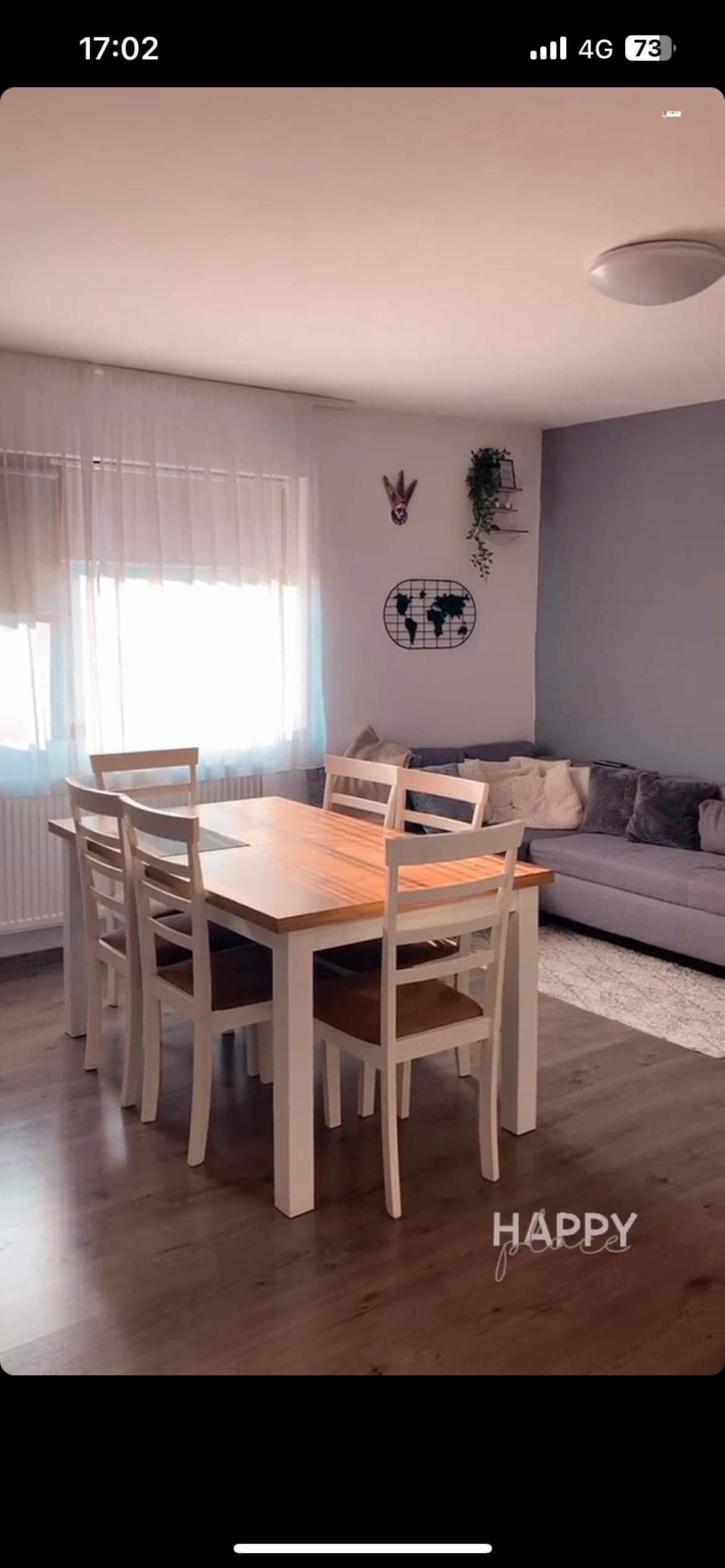 A dining area is showcased with a wooden table surrounded by four white chairs. A light-filled living space features a gray sofa adorned with pillows. Soft curtains frame the window, and wall decor adds visual interest.