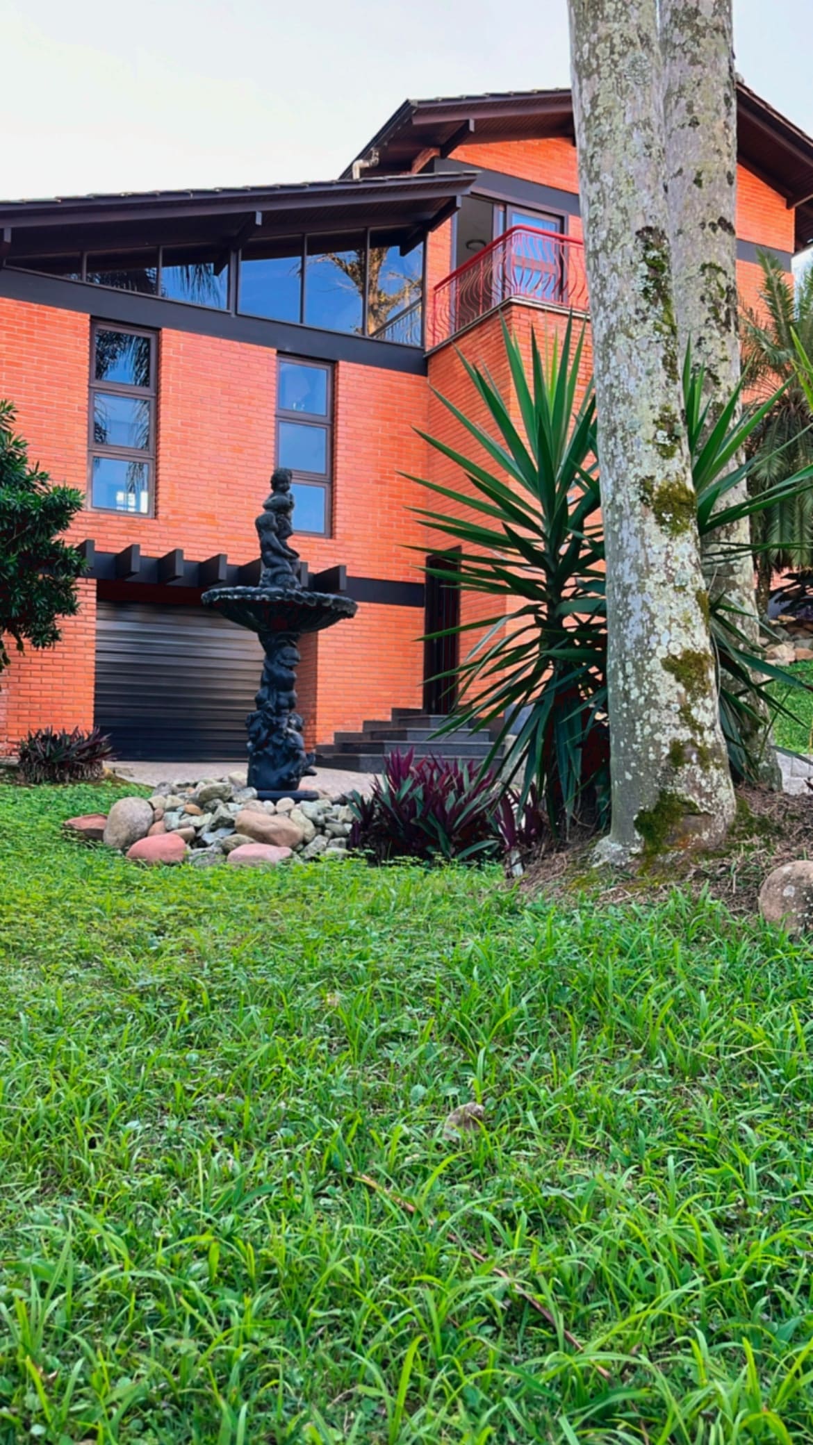 A spacious property is highlighted by a decorative fountain surrounded by a lush garden. Towering trees and vibrant plants frame the area, complementing the red brick exterior of the house, which features large windows that offer ample natural light.