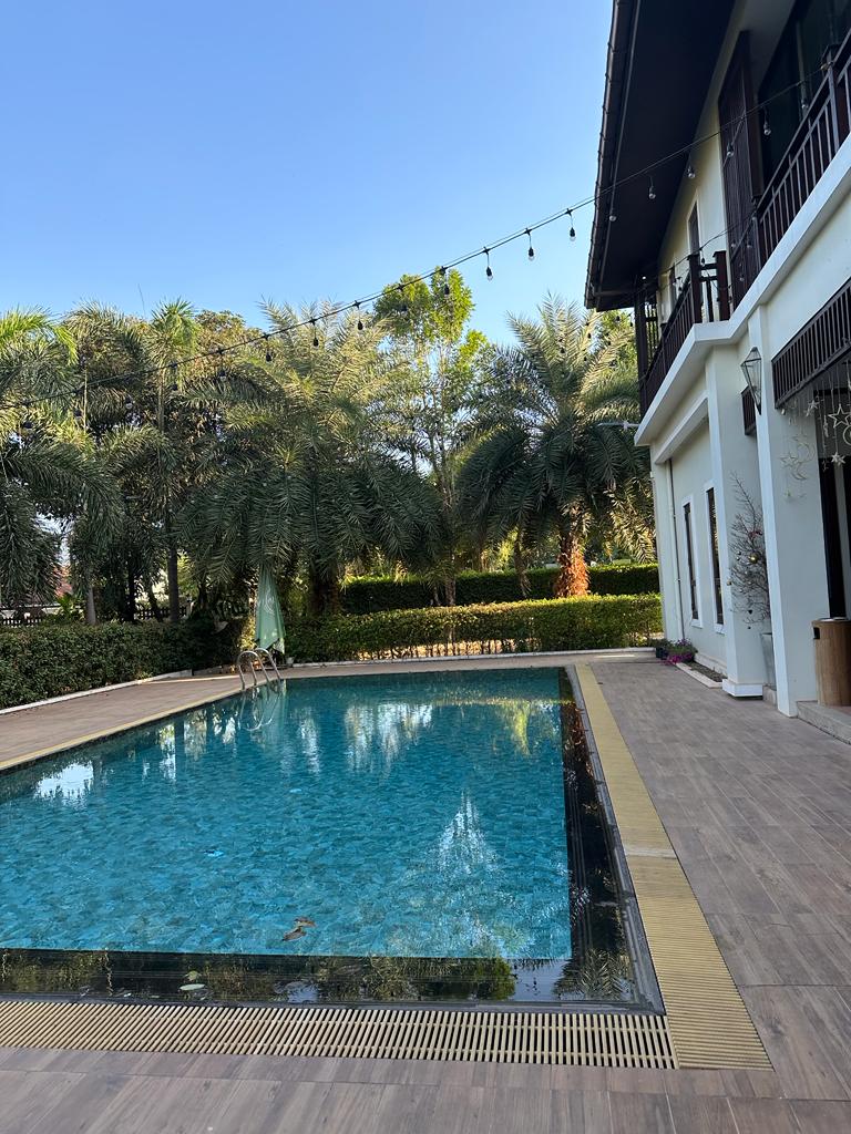 A swimming pool with clear blue water is surrounded by a tiled deck and lush greenery. Tall palm trees provide shade around the pool area. String lights are hung above, adding a subtle ambiance. The exterior of a two-story building is visible in the background.
