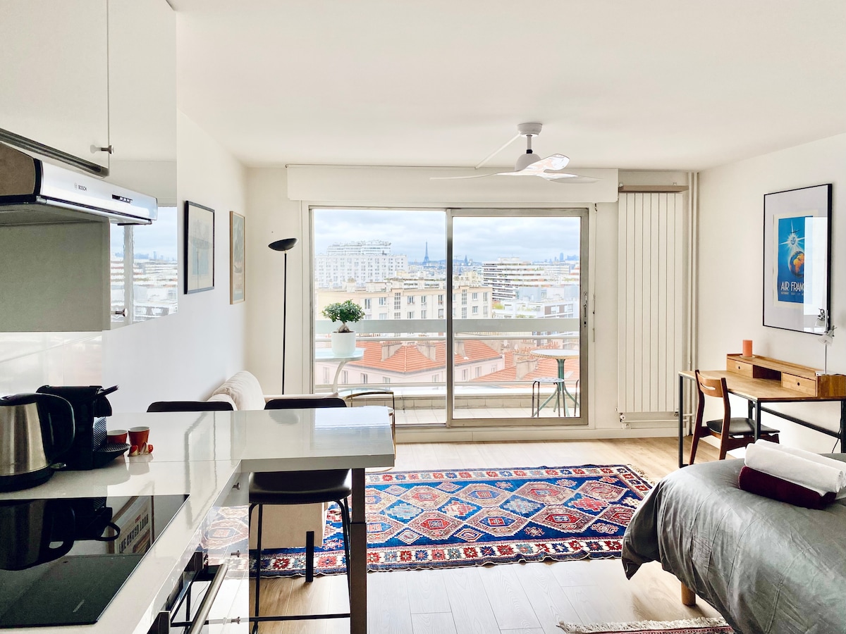 The studio layout presents a modern kitchen area with white cabinetry and a dining table. A cozy seating area is visible, complemented by a colorful rug. Large glass doors lead to a balcony, framing a panoramic view of Paris with decorative city elements in the distance.