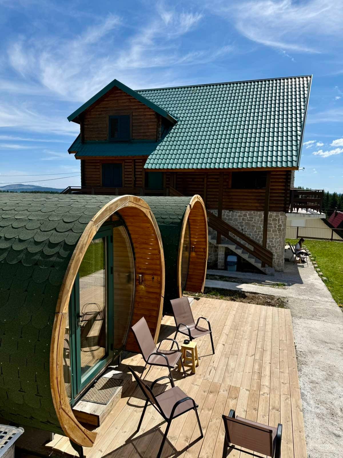 The exterior of the house is shown, featuring a traditional wooden structure with a sloped green roof. In the foreground, two round wooden pods with large glass doors are visible on a wooden deck, accompanied by multiple foldable chairs that provide an inviting outdoor space.