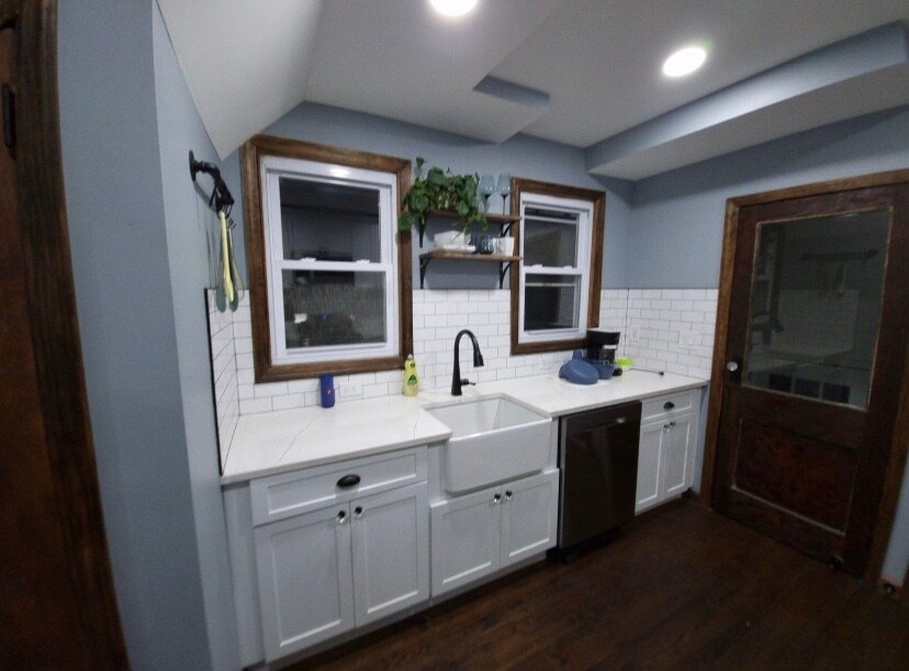 The kitchen area features sleek white cabinetry complemented by a farmhouse sink and modern faucet. A stainless steel dishwasher is integrated within the cabinetry. Natural light enters through two large windows framed in wood, enhancing the crisp and clean design of the space.
