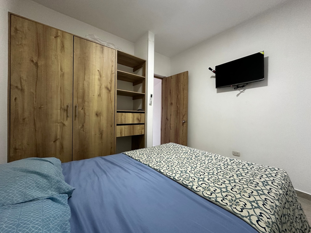 A spacious bedroom is presented with a wooden wardrobe and shelving unit. A television is mounted on the wall opposite the bed, which is dressed in a blue and white patterned bedspread. Natural light enters through the door slightly ajar, enhancing the serene ambiance.