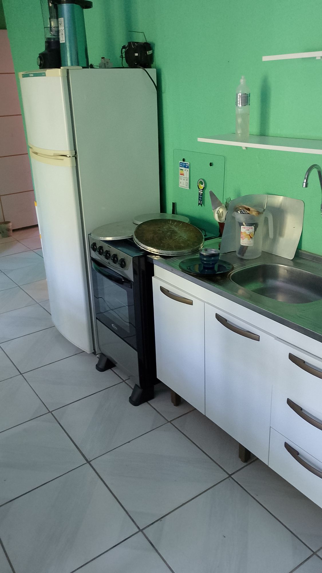 A compact kitchen space features a white refrigerator and a stove with an oven. Gray cabinets are mounted above a stainless-steel sink, with a large pan resting nearby. The green walls provide a bright backdrop, and the tiled floor is in neutral tones.