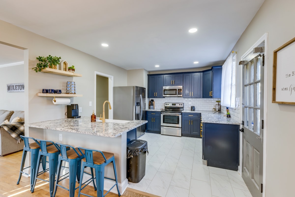 A modern kitchen features navy blue cabinetry contrasted by white countertops and stainless steel appliances. Bar stools are positioned at a spacious island, offering a communal space for dining. Natural light filters through a window above the sink, illuminating the open layout.