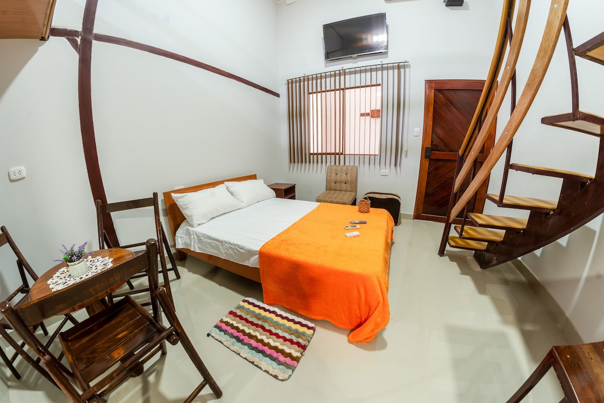 A bright room features a spacious bed covered with an orange blanket, complemented by a light-colored sheet. An intimate dining area with wooden chairs and a small table is visible. A cozy armchair is placed beside the bed, and a spiral staircase leads to an upper level.