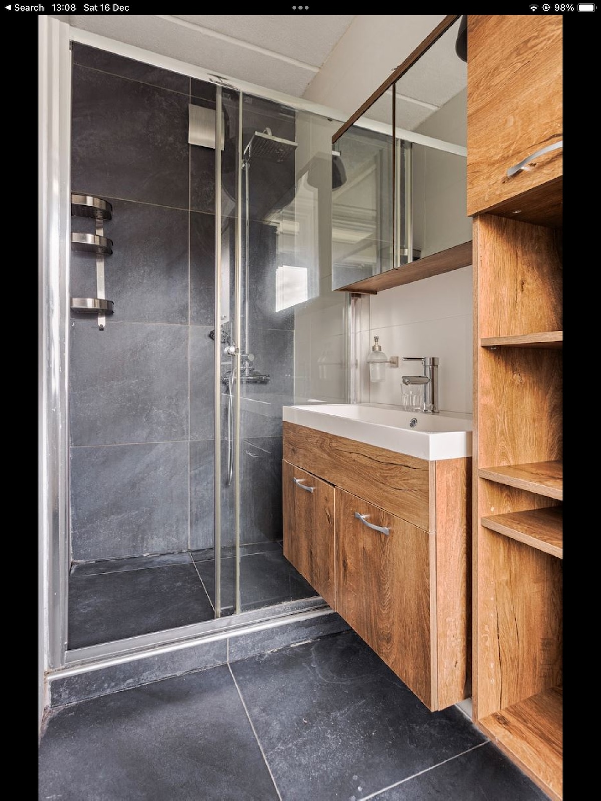 A modern bathroom features a glass shower enclosure with sleek fixtures. A wooden vanity with two cabinets and a white sink matches the overall contemporary design. The floor is covered with dark tiles, and a large mirror reflects the light-filled space.