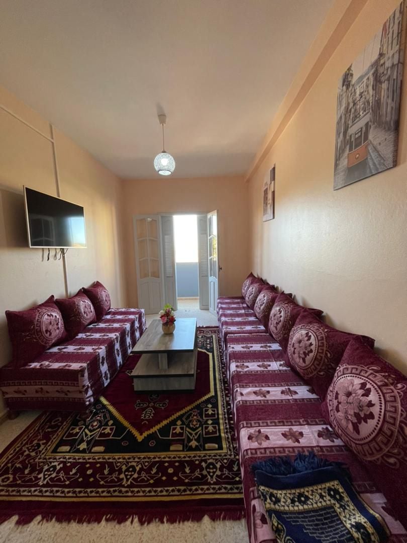 A comfortable sitting area features vibrant, red patterned cushions arranged along the walls. A low wooden table sits centrally on a decorative rug. Natural light enters through the open double doors, illuminating the light-colored walls.