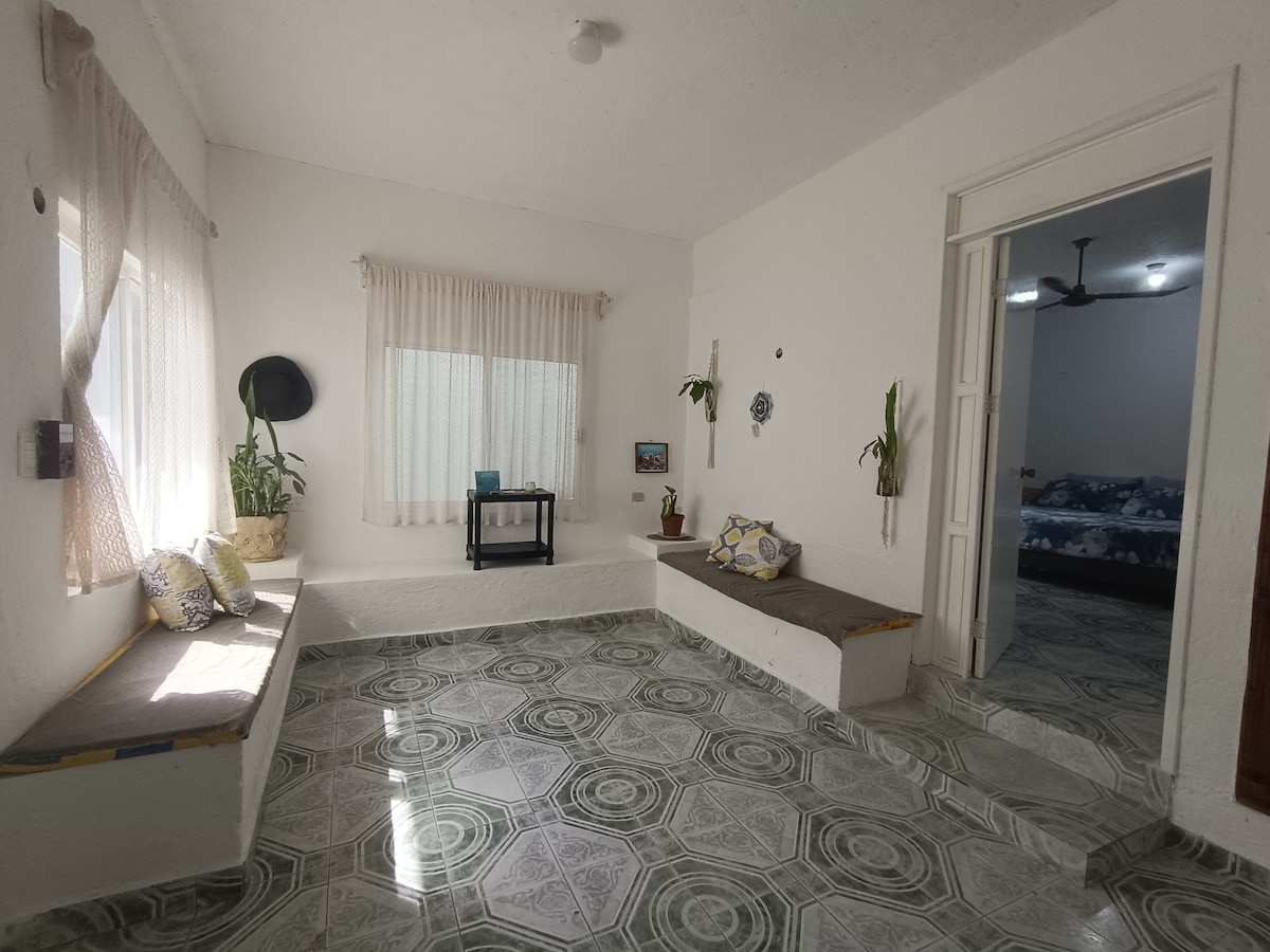 A bright and airy living space features light-colored walls and patterned tile flooring. Two built-in benches provide seating by the windows, adorned with potted plants. Natural light filters through sheer curtains, illuminating the room, which opens to a bedroom area in the background.