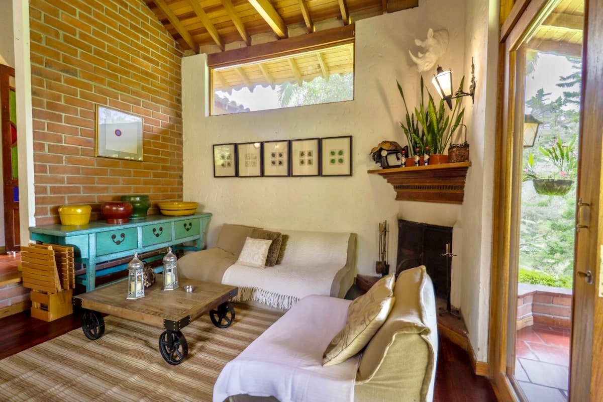 A cozy living area features a combination of light and dark furnishings. A rustic coffee table sits atop a wooden floor, accompanied by two sofas draped in neutral tones. Artwork hangs on the wall above a brightly colored sideboard, and natural light enters through large windows.