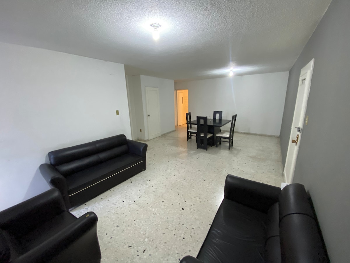 A spacious living area is furnished with two black sofas and a six-seat dining table. The walls are painted in light colors, enhancing the bright atmosphere, while the textured flooring adds depth to the room. A door leads to another area of the apartment.