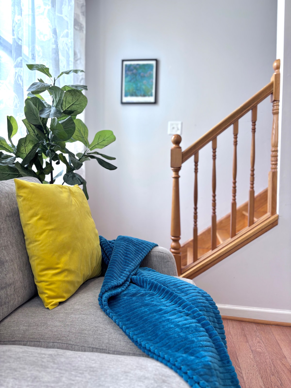A cozy seating area is highlighted by a gray sofa adorned with a vibrant yellow pillow and a blue throw blanket. A leafy plant enhances the ambiance near the window, while a staircase with wooden railings leads to the upper level. A framed artwork adds subtle color to the wall.