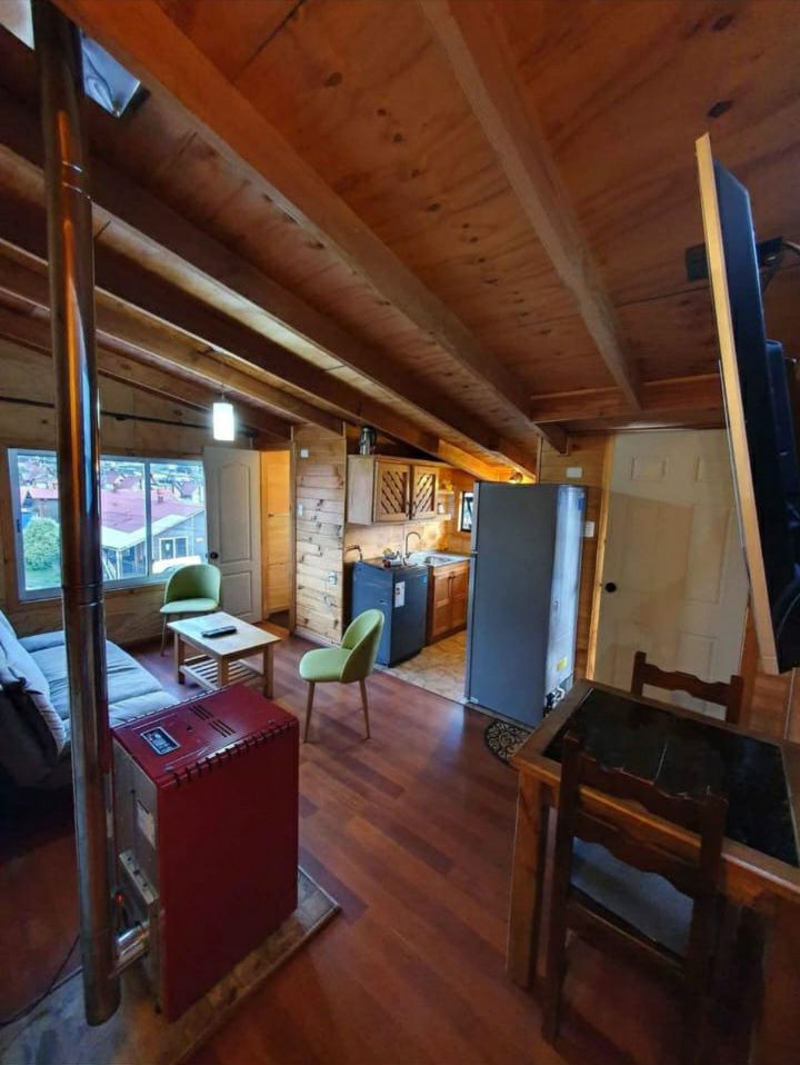 The interior space features wooden walls and a high ceiling with exposed beams. A compact kitchen is visible, alongside a small dining table and two chairs. A cozy seating area includes a green chair and a wood-burning stove, contributing to the warm ambiance.