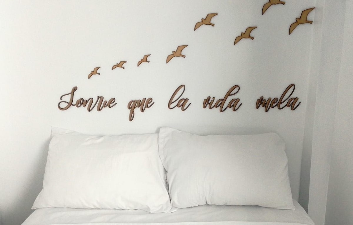 A spacious bed is positioned against a white wall, adorned with wooden decor depicting birds in flight. Above the bed, a wooden phrase in Spanish reads 'Sonríe que la vida vuela,' complementing the serene atmosphere.
