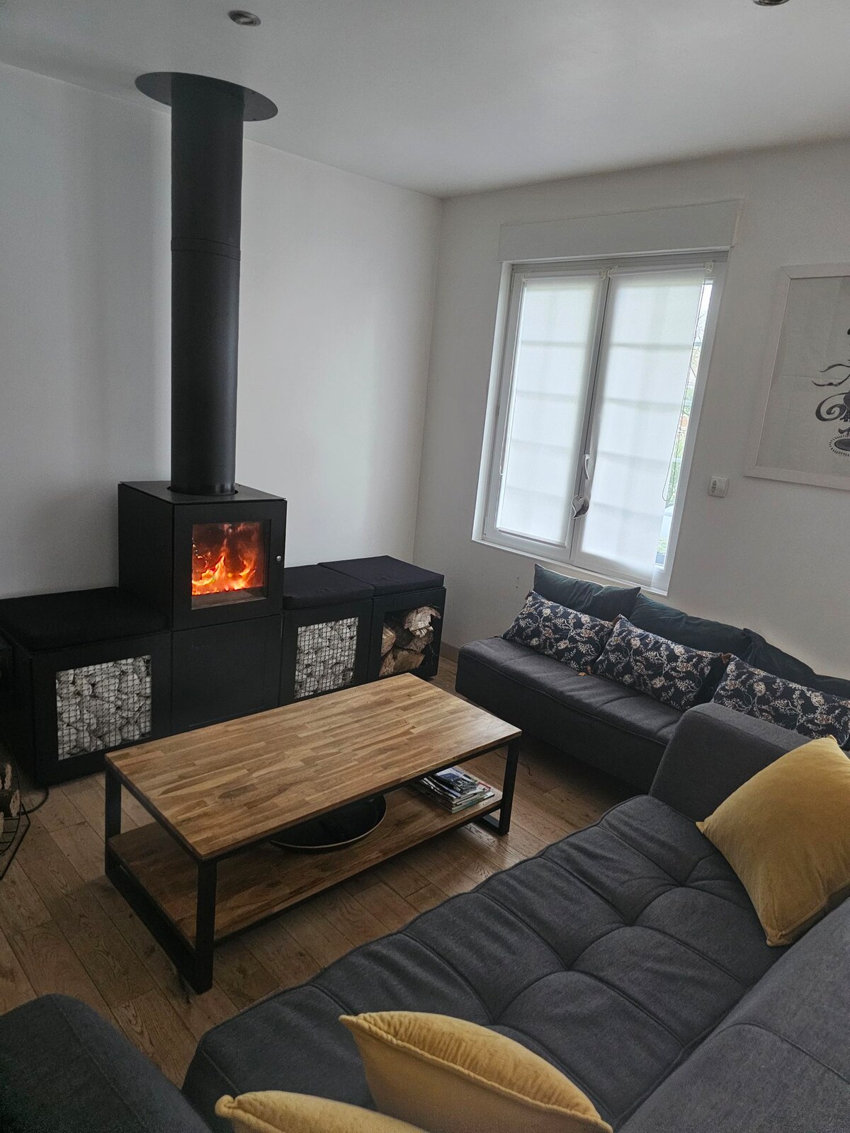 A cozy living area is highlighted by a modern wood stove, offering warmth. A comfortable gray sofa adorned with yellow cushions faces a wooden coffee table, while natural light enters through a large window with double panes, enhancing the welcoming ambiance.