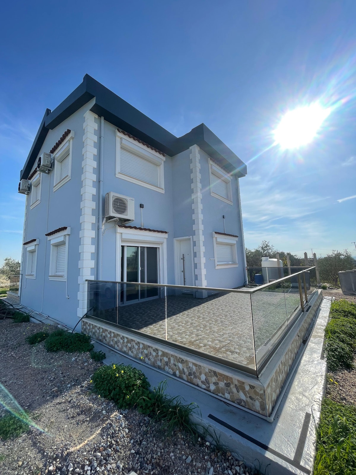 A modern two-story house is depicted, featuring a blue exterior and a glass-railed terrace. The structure is accompanied by several windows and air conditioning units. Sunlight creates a bright, clear atmosphere, while surrounding greenery adds a natural element to the setting.