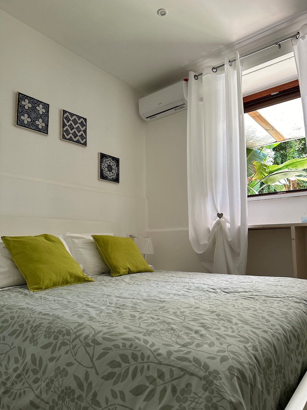 The sleeping area features a spacious bed adorned with a light floral patterned coverlet and two green decorative pillows. Soft natural light enters through a window, partially covered by sheer white curtains. Wall art complements the serene atmosphere, while an air conditioning unit offers comfort.