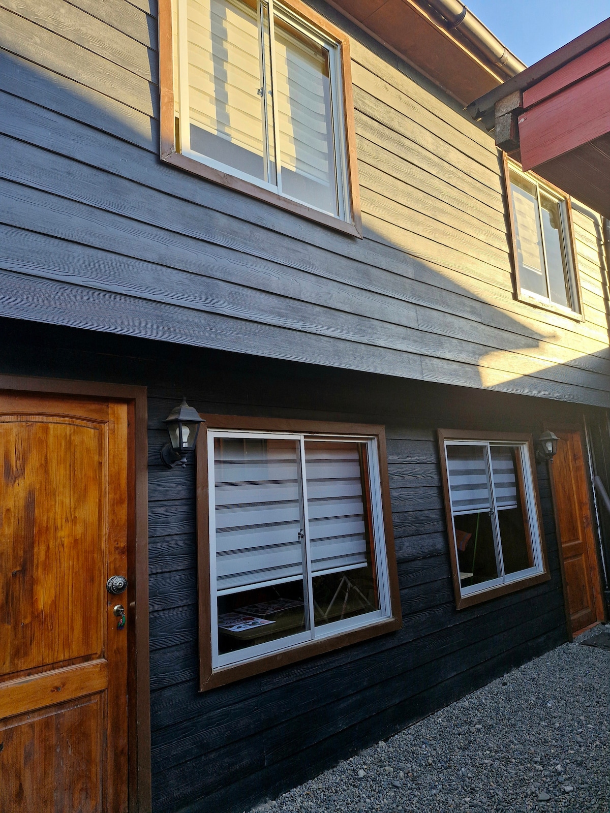The exterior of the cabin is shown, featuring a dark wooden facade with large windows framed in light wood. The entrance door is wooden with a round doorknob, and wall-mounted outdoor lights are positioned beside the windows.
