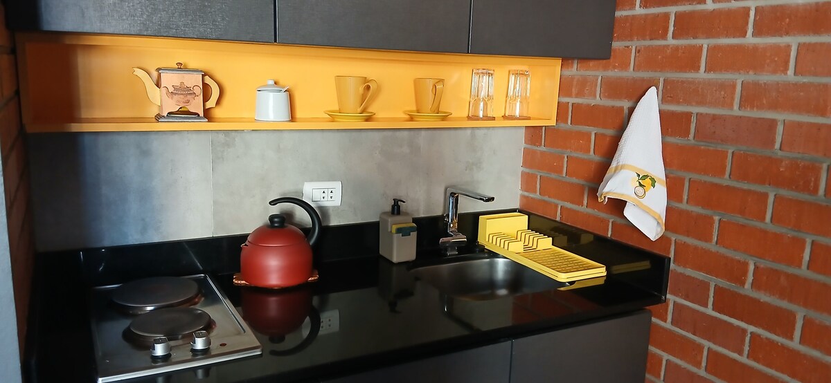 A compact kitchen area features dark cabinetry paired with a vibrant orange shelf. An electric stovetop and a sleek black sink are visible, along with a red kettle and a collection of yellow and clear glass utensils arranged neatly. A white towel hangs beside the sink.