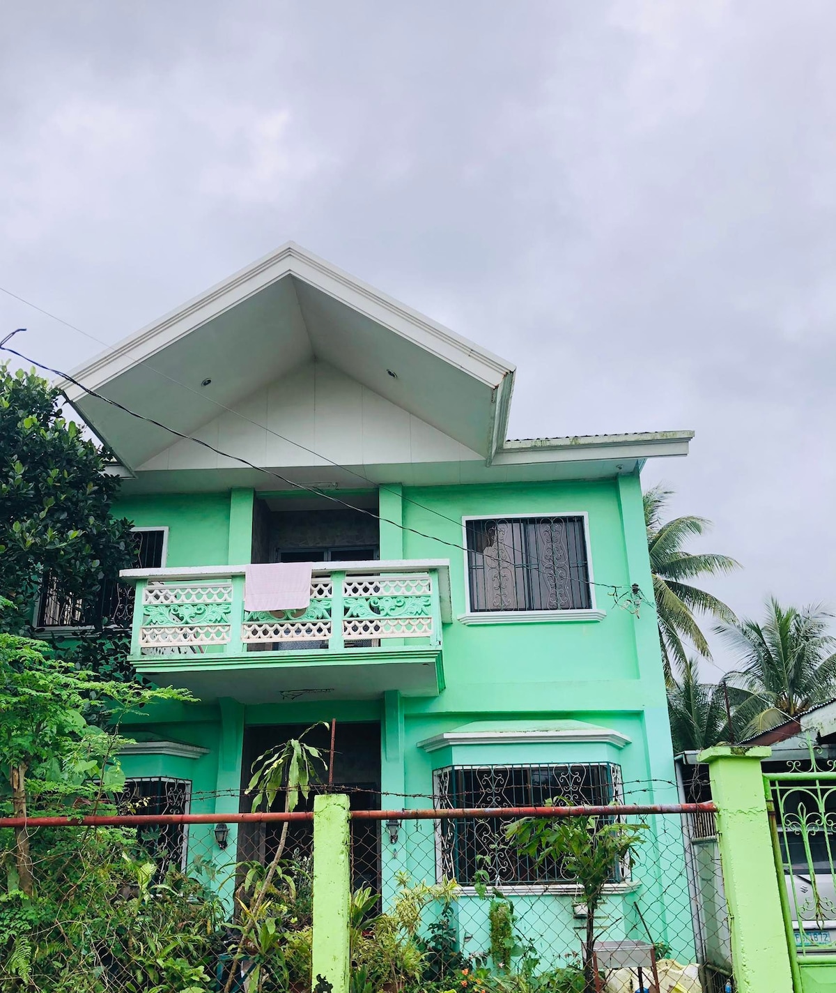 A two-story house is presented with a distinct green exterior and a sloping roof. The second floor features a balcony with decorative railings, while large windows offer views of lush surrounding greenery. A fence encircles the property, enhancing its privacy.