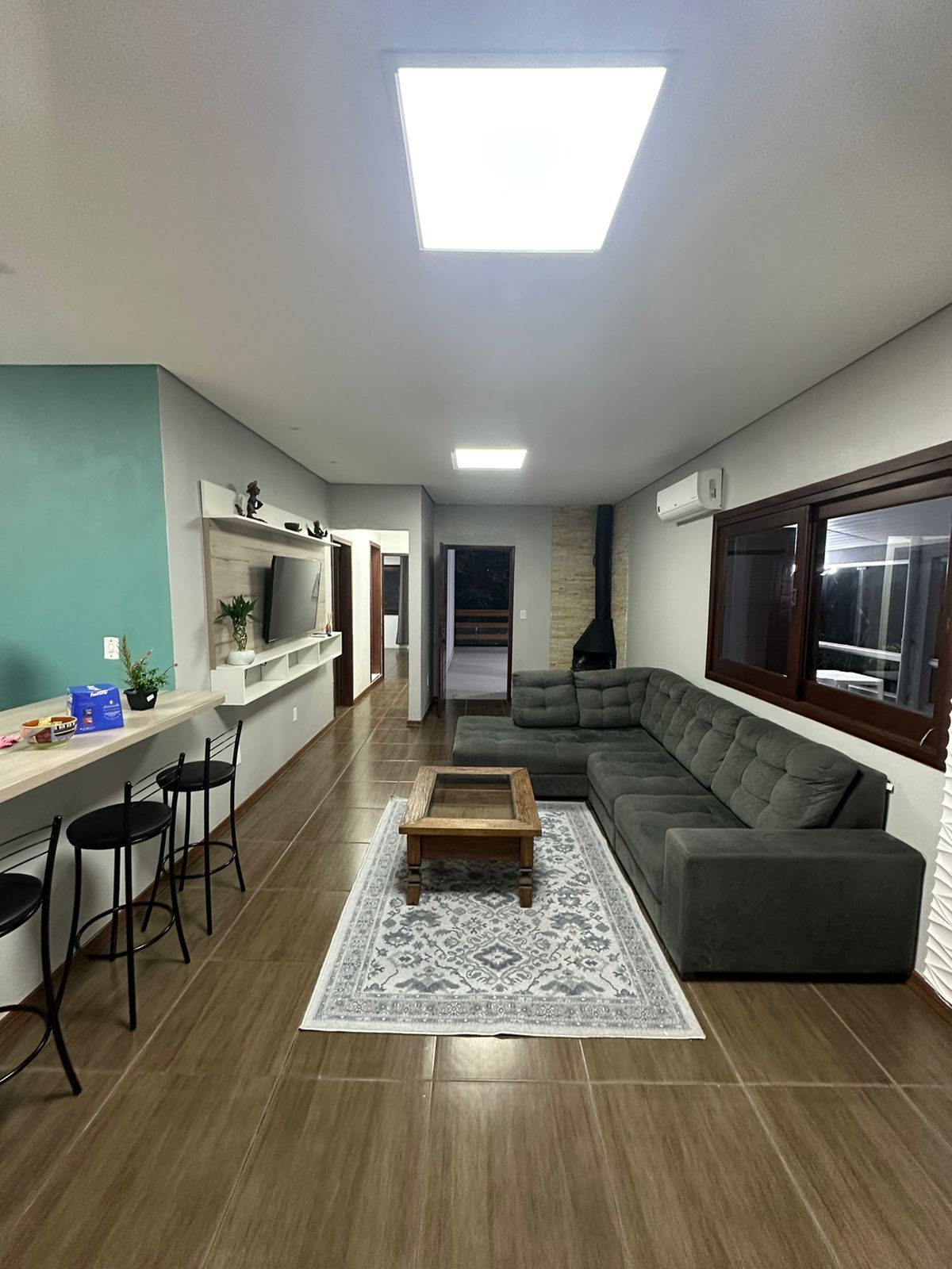A spacious living area features a large sectional sofa in a muted tone, paired with a wooden coffee table atop a patterned area rug. Large windows allow for natural light, while wall-mounted shelves and a flat-screen TV complete the inviting space.