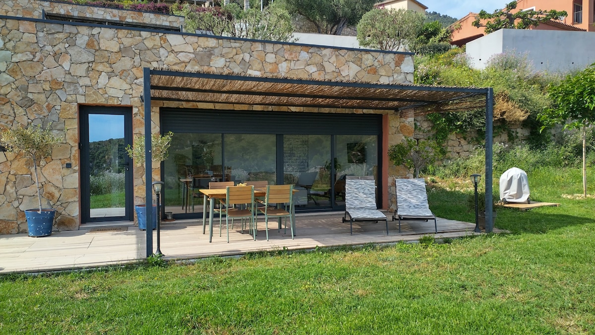 An outdoor terrace features a dining table surrounded by four chairs, and two lounge chairs, all situated on a grassy area. Large glass doors provide a view into the interior space. The terrace is framed by a pergola, with surrounding greenery enhancing the serene setting.