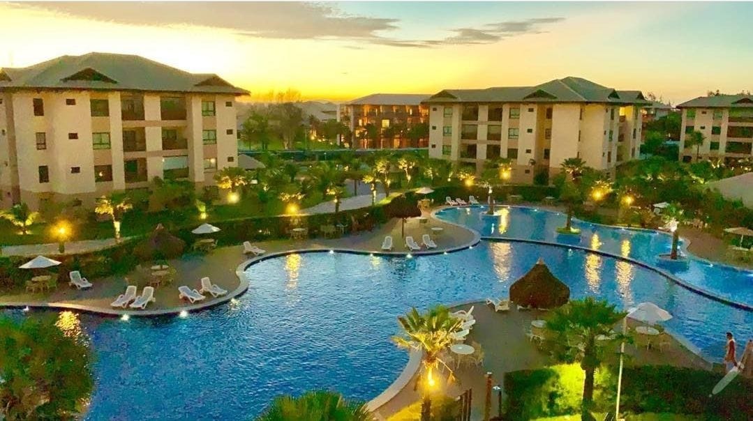 A well-lit outdoor space features an expansive swimming pool surrounded by palm trees and lounge chairs. Elegant buildings are visible in the background against a sunset sky, creating a calm and relaxed ambiance.