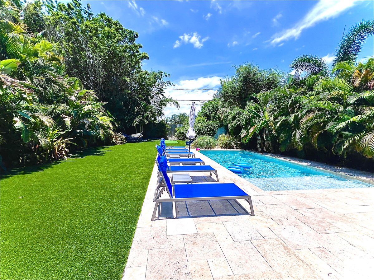 A well-maintained outdoor area features a heated pool surrounded by tropical landscaping. Six blue lounge chairs are positioned on the sunlit stone patio, offering a space for relaxation. Lush greenery provides privacy, while an umbrella stands near the pool for shade.