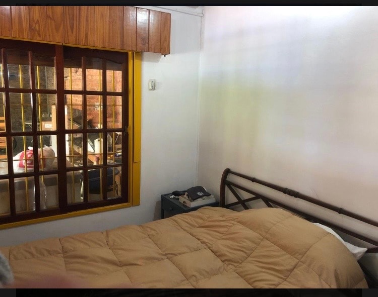 A cozy sleeping area featuring a spacious bed covered with a soft, neutral-toned blanket. A wooden headboard complements the design. Natural light enters through a window adorned with a grid of mullions, providing a glimpse of the vibrant outdoor setting.