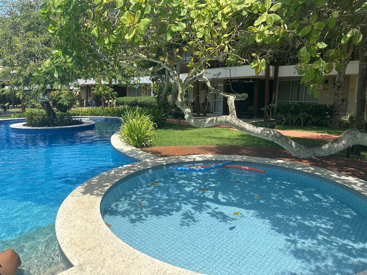 An inviting pool area is surrounded by lush greenery, with a larger swimming pool and a smaller spa-style pool. A large tree provides shade over the patio, enhancing the tranquil atmosphere. A walkway is visible, connecting to nearby accommodations.