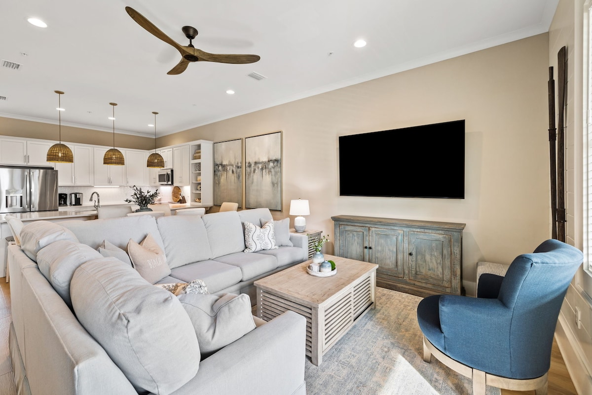 An open living space features a pale blue sectional sofa with decorative pillows, positioned around a wooden coffee table. A large flat-screen TV is mounted on the wall, and light oak cabinetry is visible in the background, blending seamlessly with the light walls.