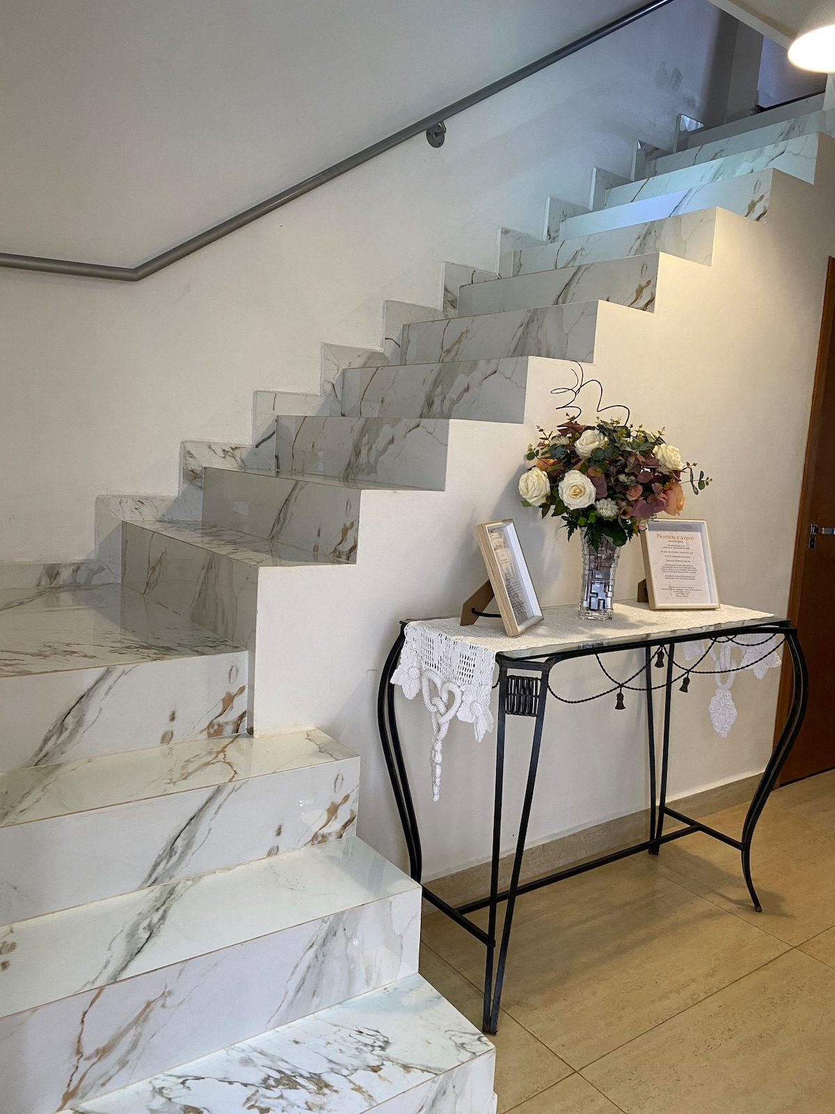 A modern staircase features marble steps, leading to an upper level. A decorative console table is positioned against the wall, adorned with a floral arrangement and framed documents. Soft lighting enhances the elegant atmosphere of the entryway.