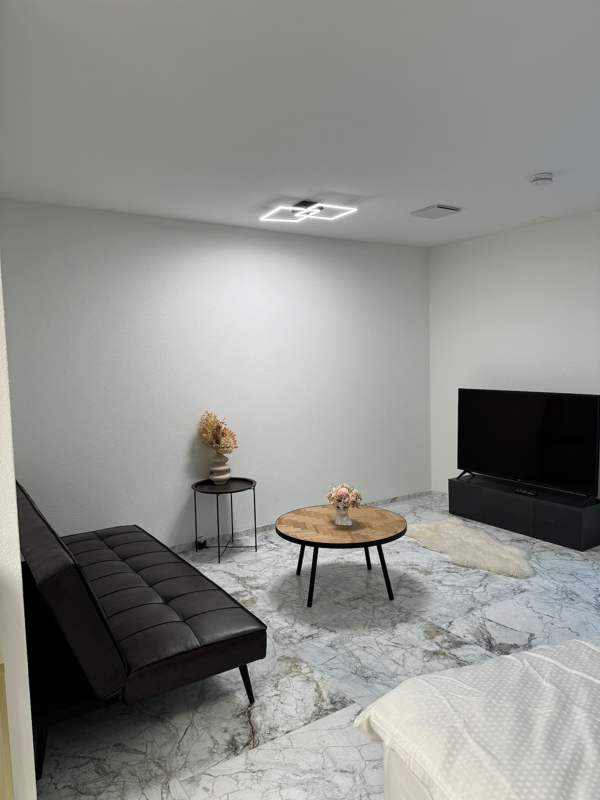 A seating area is featured with a dark sofa positioned near a round wooden coffee table. A small vase holds dried plants on a nearby side table. The space is complemented by a television against a light wall, with patterned tile flooring adding texture.