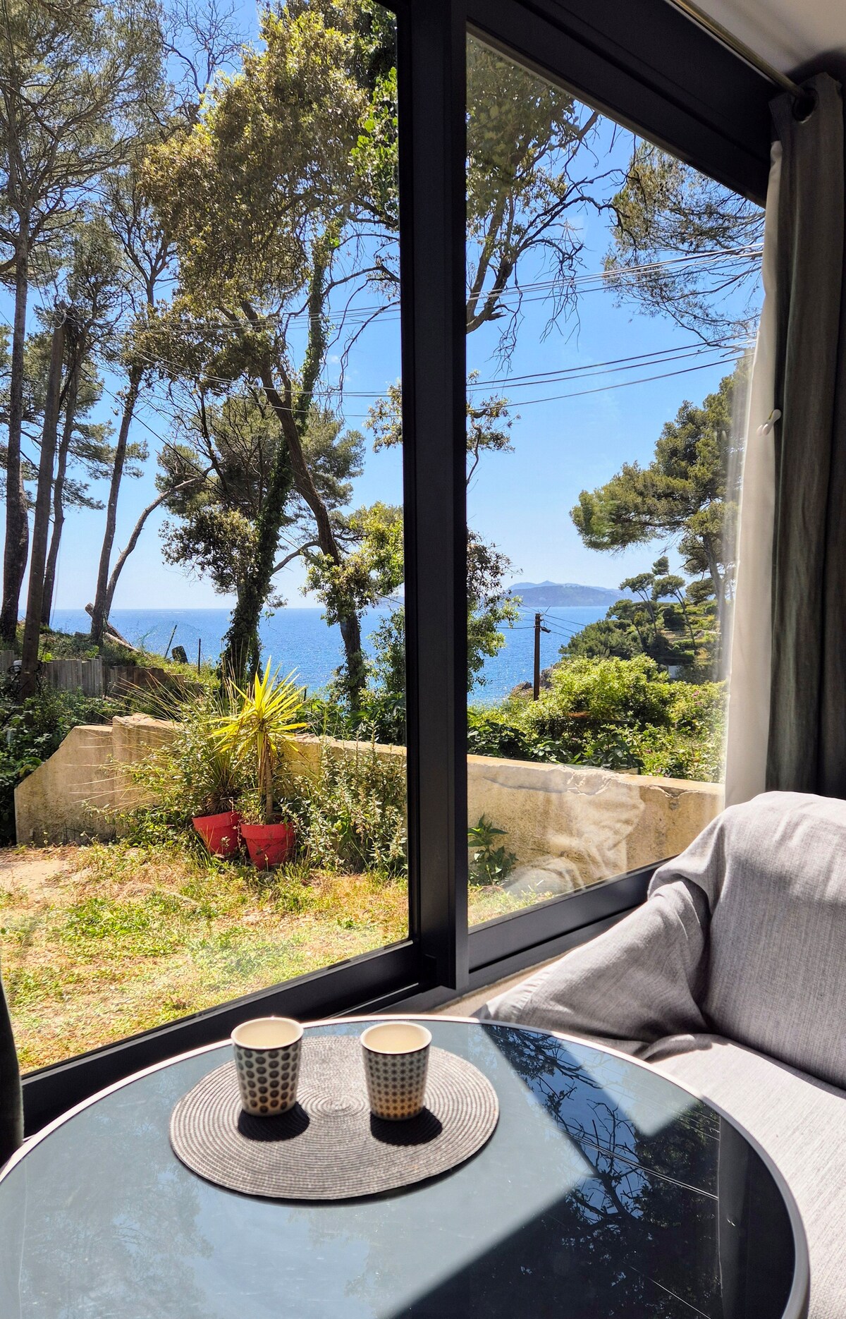 A cozy seating area with a view through large glass windows showcases a blend of indoor comfort and outdoor tranquility. A small, round table holds two cups, and sunlight illuminates the space, while lush greenery and glimpses of the sea can be seen in the background.
