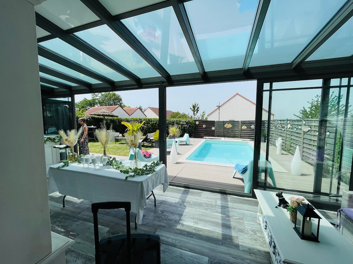 A spacious veranda features large glass walls, providing a clear view of the pool and garden. A beautifully arranged dining table with decorative elements sits within, enhancing the inviting atmosphere. Sunlight filters through the glass roof, illuminating the modern decor.