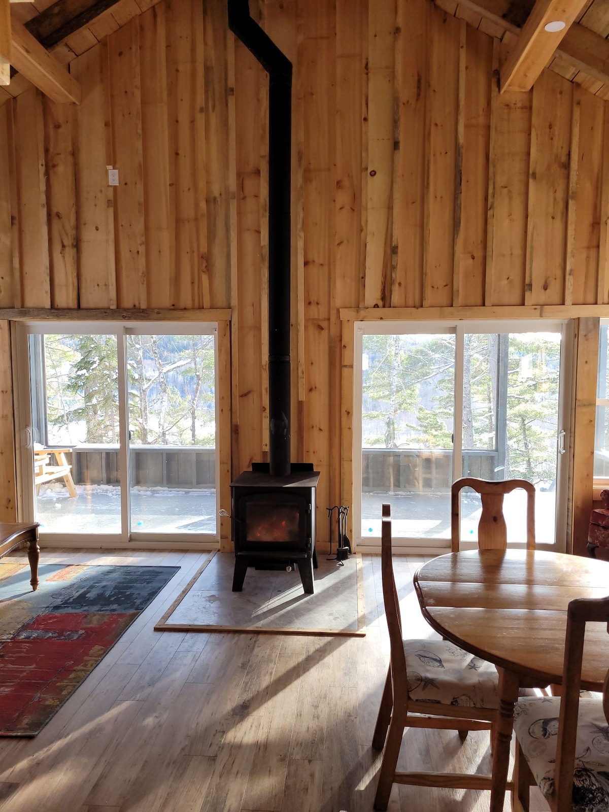 Tall wooden walls create a warm ambiance around a wood stove, positioned centrally within the room. Light streams through multiple glass doors, illuminating a small dining table with chairs, while a cozy seating area can be seen nearby.