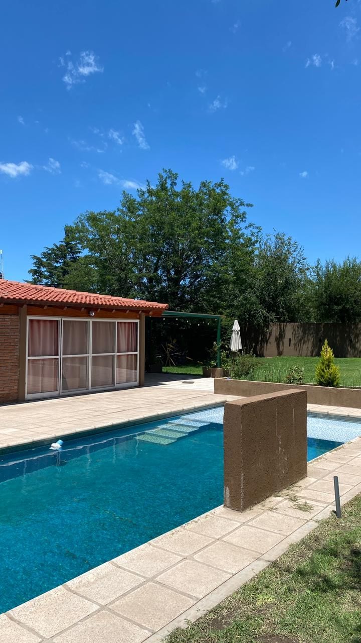A well-maintained outdoor area features a clear swimming pool surrounded by a stone patio. Lush greenery is present in the background, with a sun umbrella positioned on the lawn. A brick structure with large glass doors completes the inviting space.