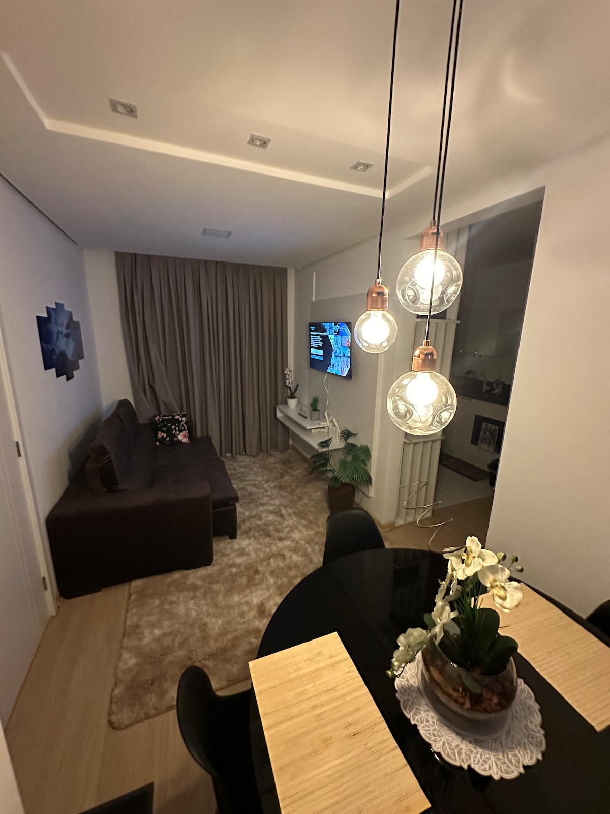 A cozy living area is arranged with a dark-colored sofa and a flat-screen television mounted on the wall. Soft lighting is provided by pendant fixtures above a round dining table, which features a floral centerpiece. A plush area rug adds comfort to the space.