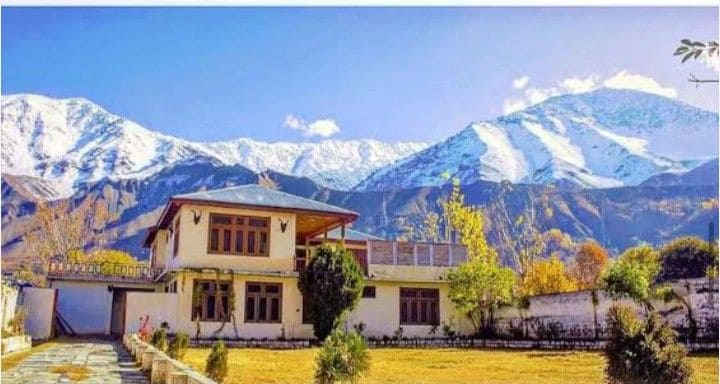 A spacious building is set against a backdrop of snow-capped mountains, surrounded by a landscaped yard. Large windows allow views of the scenic landscape. Lush greenery is present in the garden, complementing the tranquil environment of this mountain escape.