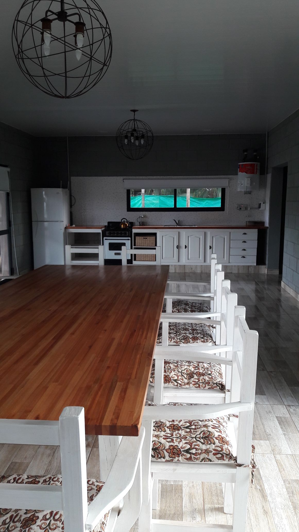 A spacious kitchen is presented, featuring a long wooden dining table surrounded by several white chairs. Modern appliances, including a refrigerator and stove, are visible against a neutral wall, while a large window allows natural light to illuminate the area.