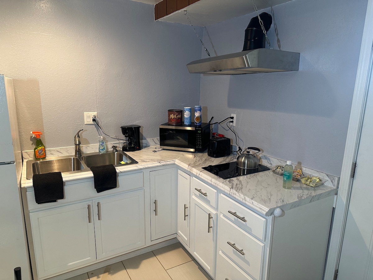 A renovated kitchen is presented with white cabinetry and a marble countertop. Two sinks are positioned side by side, with essential appliances such as a microwave, coffee maker, and kettle visible. Kitchen towels are neatly placed on the countertop, contributing to an organized and functional space.