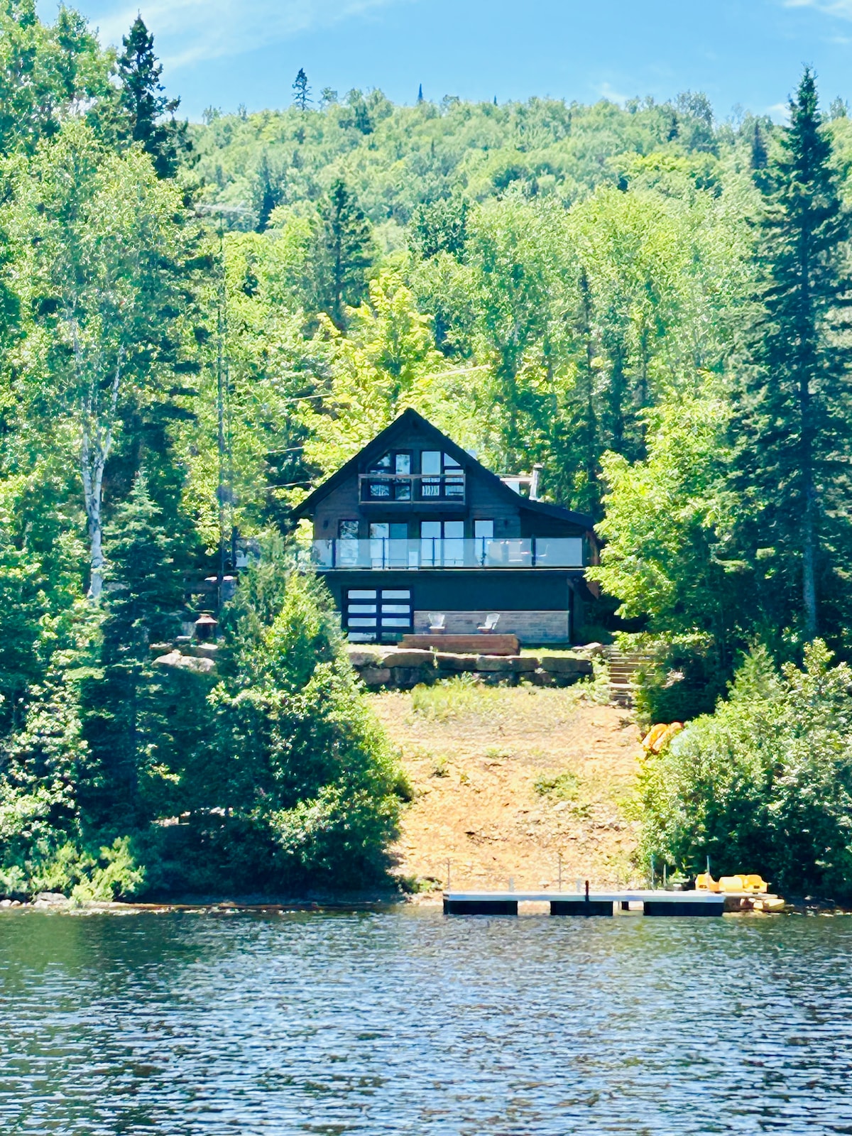 A wooden chalet is positioned on a hillside, surrounded by lush greenery and trees. The building features a mix of dark and light tones and has large windows. A dock can be seen at the lakeshore, with a hint of the tranquil water's edge below.