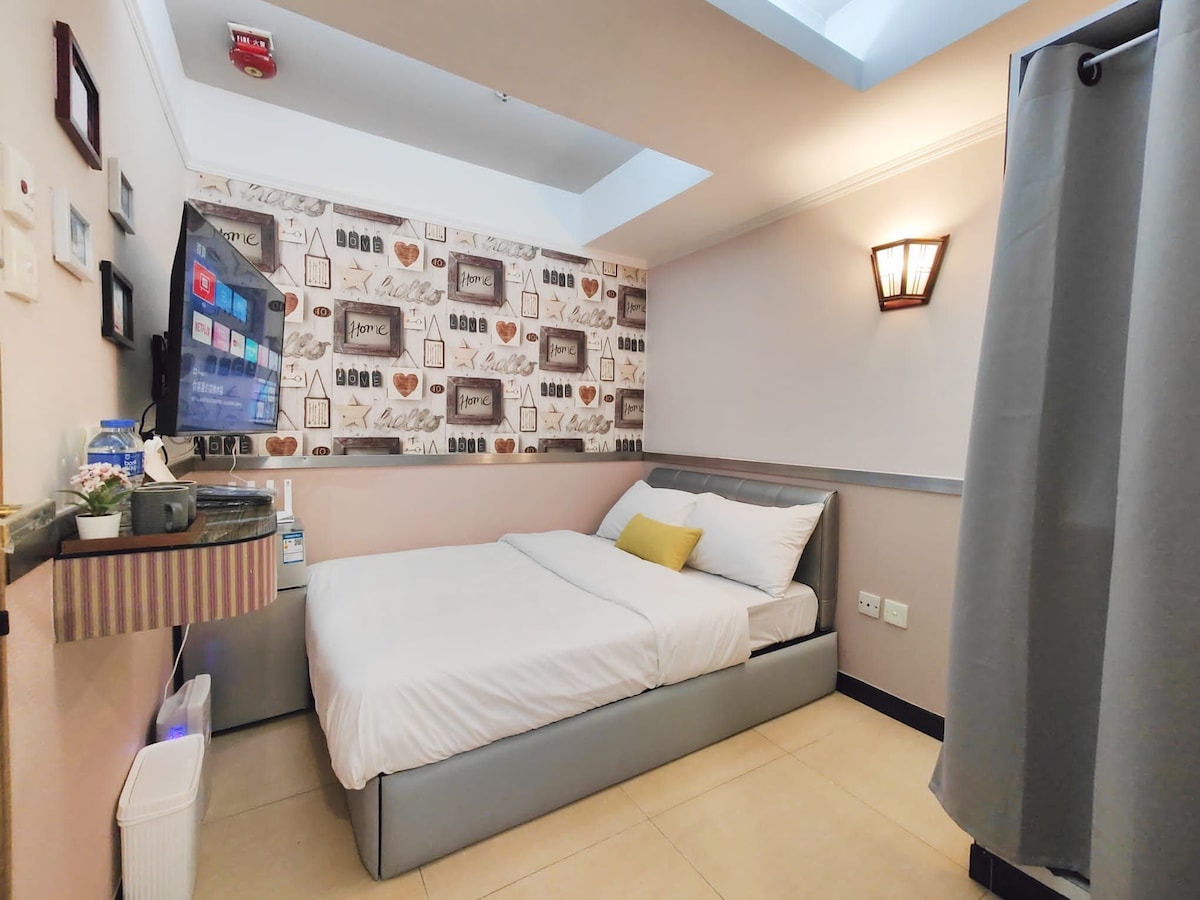 A minimalist bedroom features a spacious bed adorned with crisp linens. A small decorative pillow adds a pop of color. A television is mounted on the wall, and a window above allows natural light to brighten the space. Curved shower curtains provide privacy.