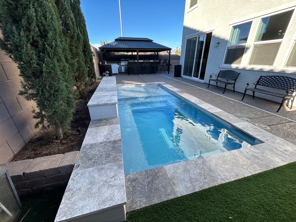 A rectangular swimming pool is framed with light-colored stone tiles, reflecting the clear blue sky. A shaded seating area with black benches is visible nearby, offering a relaxed space for gatherings. Lush greenery stands along the perimeter, enhancing the outdoor view.
