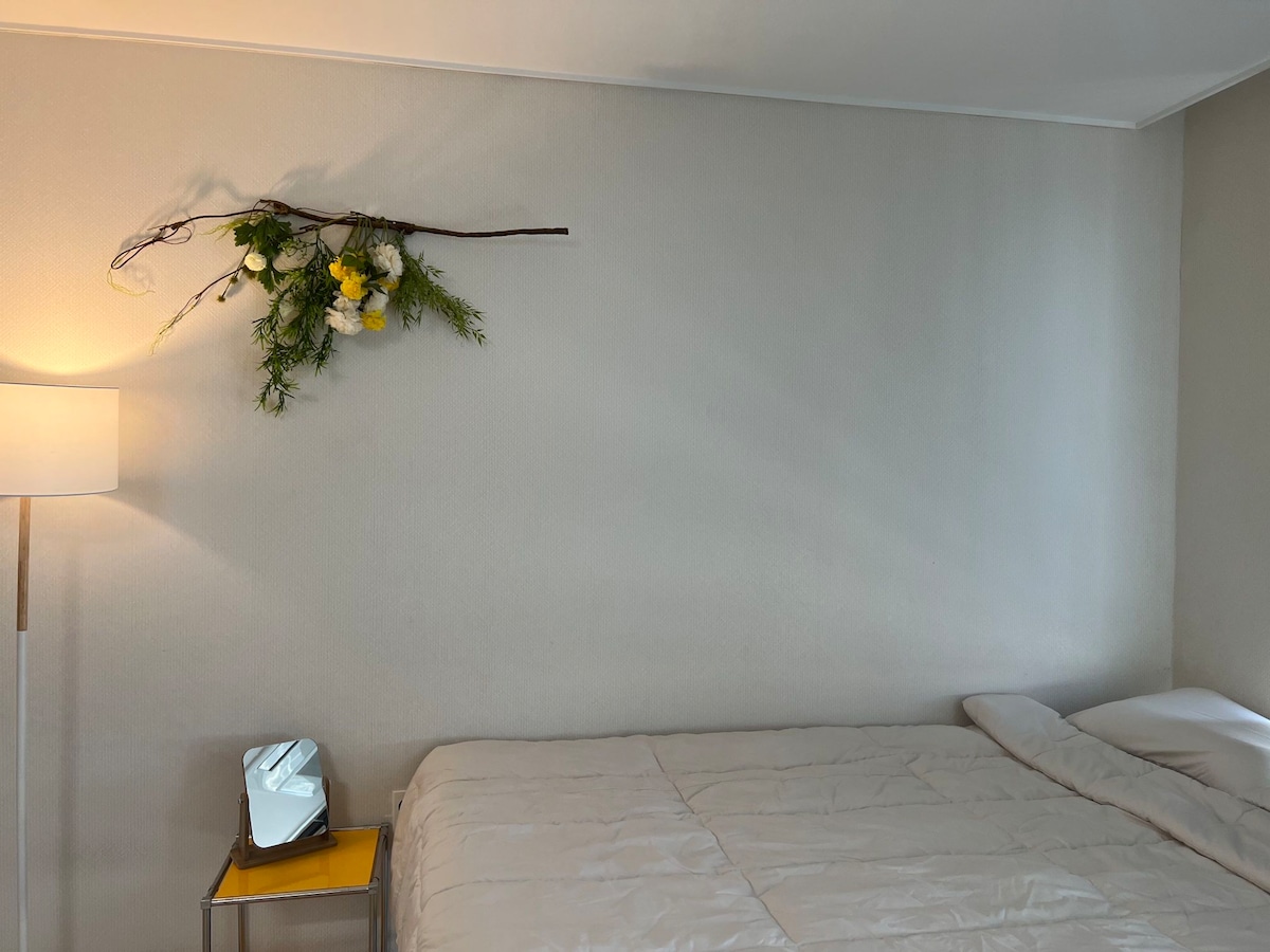 A minimalist bedroom features a spacious bed with a light-colored coverlet. A small mirror and a yellow side table are positioned beside the bed. Above, a decorative branch adorned with flowers and greenery adds a touch of nature to the neutral wall.